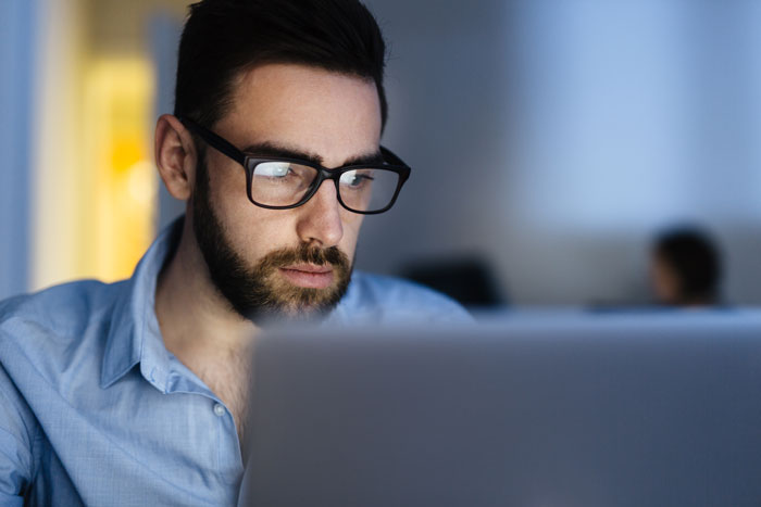 young male with beard professional in an office wearing glasses working and focusing on laptop