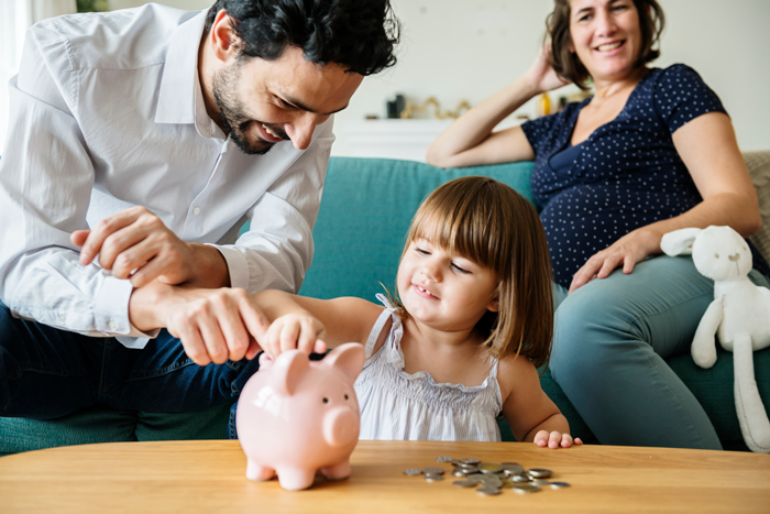 parents watching and smiling at little girl putting coins in a piggy bank